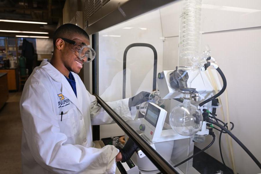 Male UTC Chemistry student wearing safety goggles and a lab coat standing next to lab equipment
